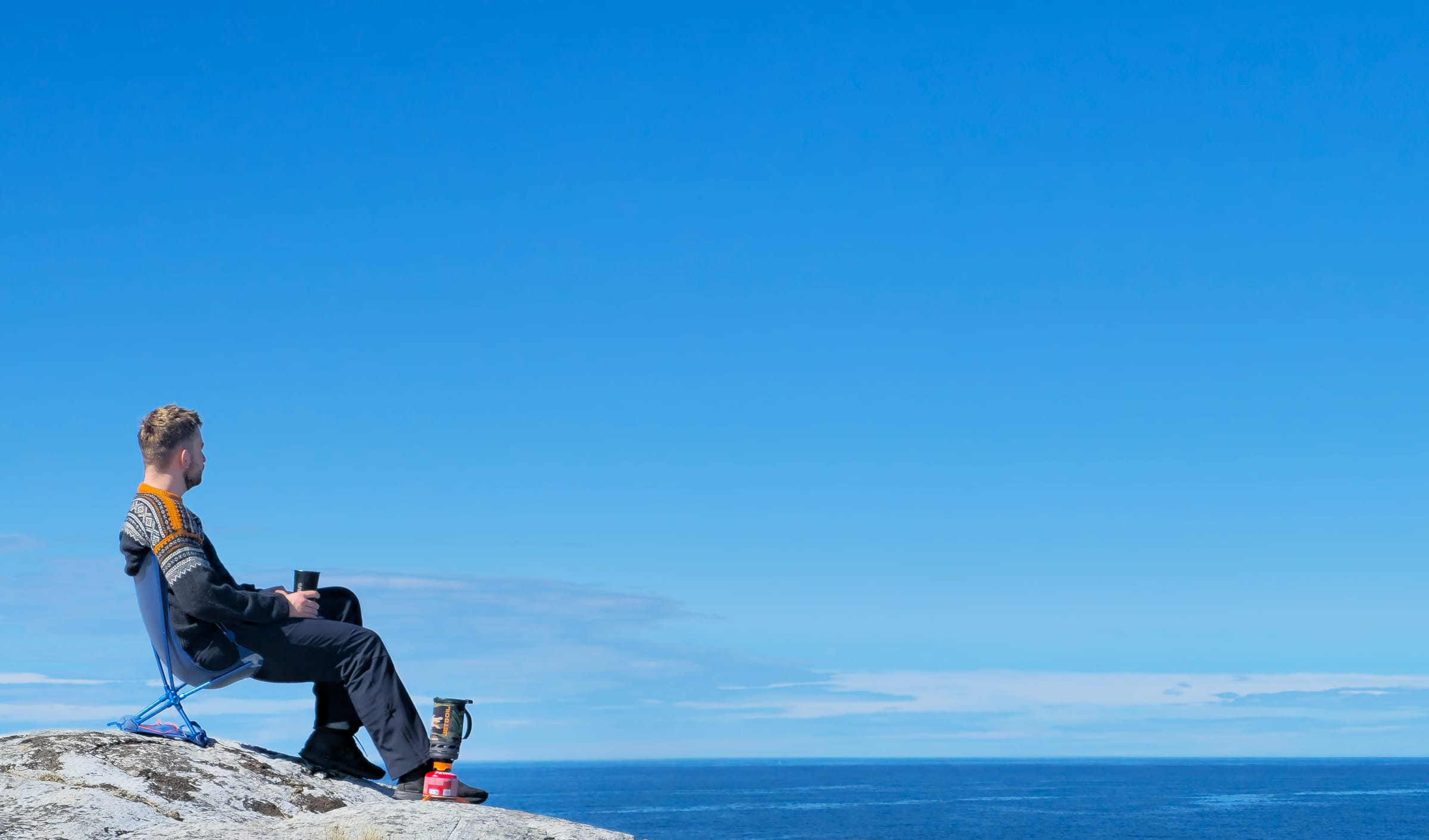 Turgåer i mariusgenser sitter i turstol på svaberg ved havet med kaffekopp og turbrenner